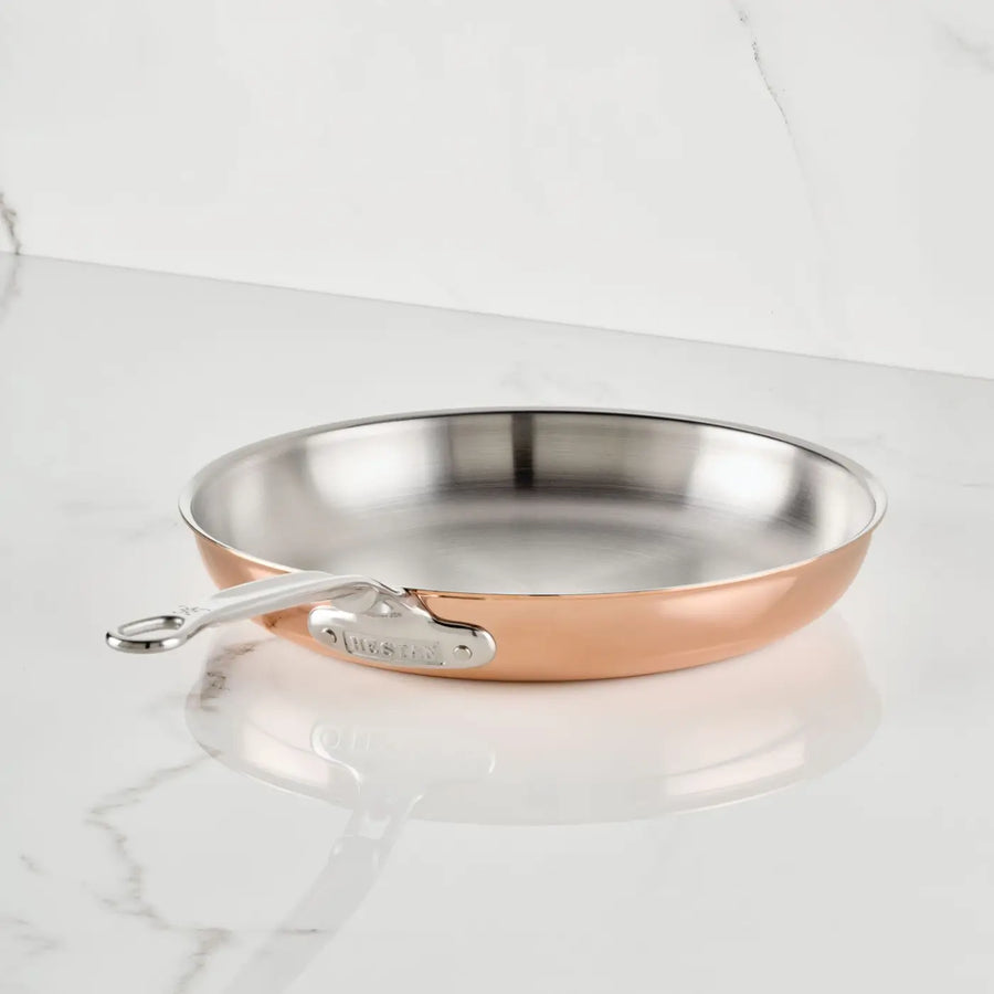 A Polished Copper Skillet by CopperBond Aura™ with a stainless steel interior and metal handle rests on a glossy white marble countertop against a white marble backsplash.