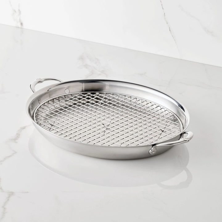 The Hestan Provisions 17.5-Inch x 14-Inch Clad Oval Roaster & Rack features dual handles and a removable wire rack, elegantly displayed on a white marble surface.