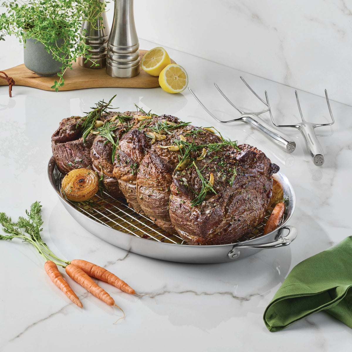 A cooked roast beef garnished with herbs sits in the Hestan Provisions 17.5-Inch x 14-Inch Clad Oval Roaster & Rack, surrounded by onions and carrots. Nearby are fresh herbs, pepper grinders, lemons, two meat forks, and a green napkin on a marble countertop.