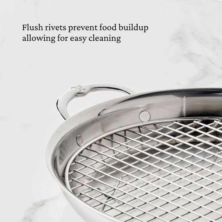 A close-up of a Hestan Provisions 17.5-Inch x 14-Inch Clad Oval Roaster & Rack showcases its stainless steel design with flush rivets and an included metal rack. Text above reads, “Flush rivets prevent food buildup allowing for easy cleaning.” A marble surface is visible in the background.