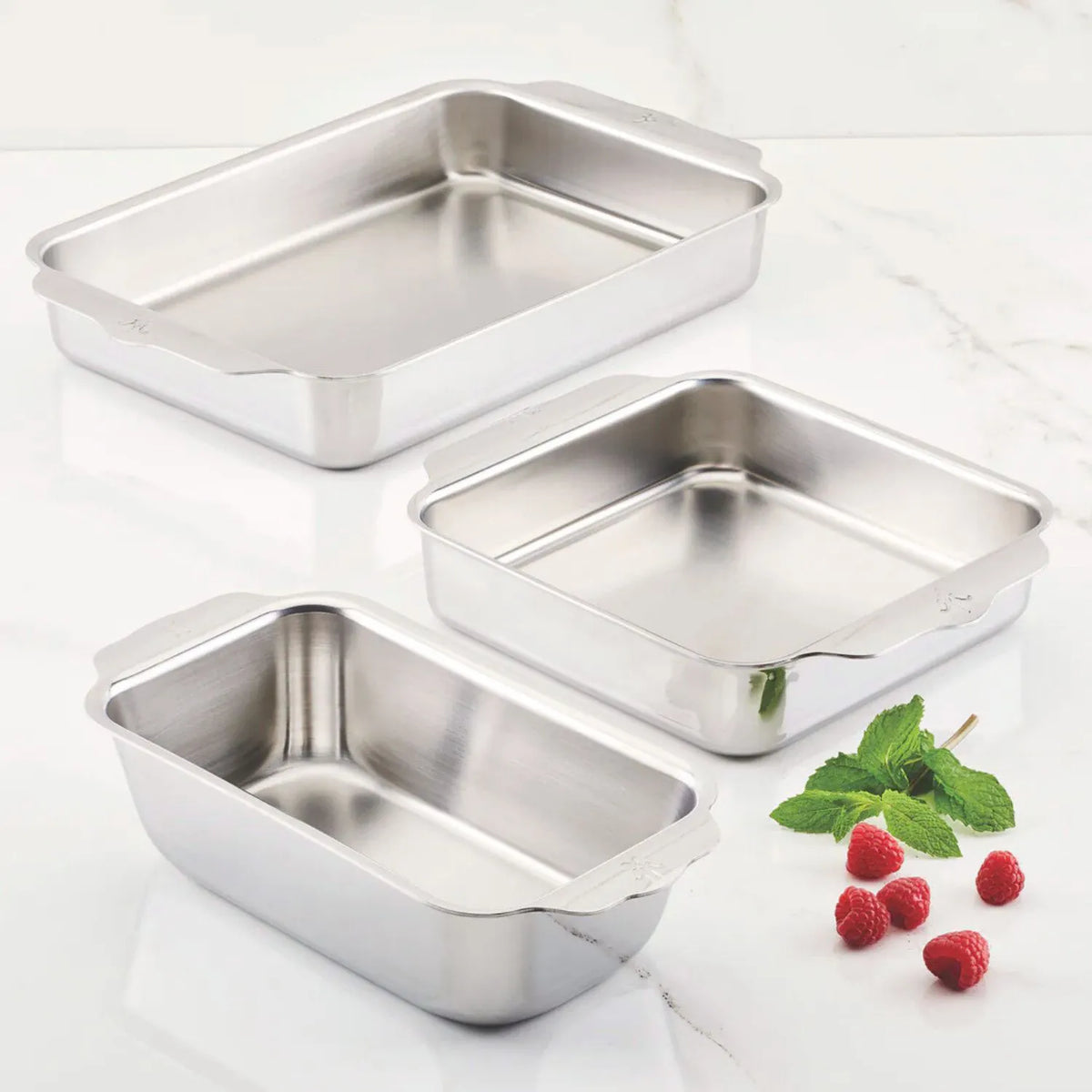 The Hestan OvenBond Tri-Ply 3-Piece Baking Set is displayed on a white marble surface, accented with raspberries and mint leaves next to this stylish, oven-safe bakeware.