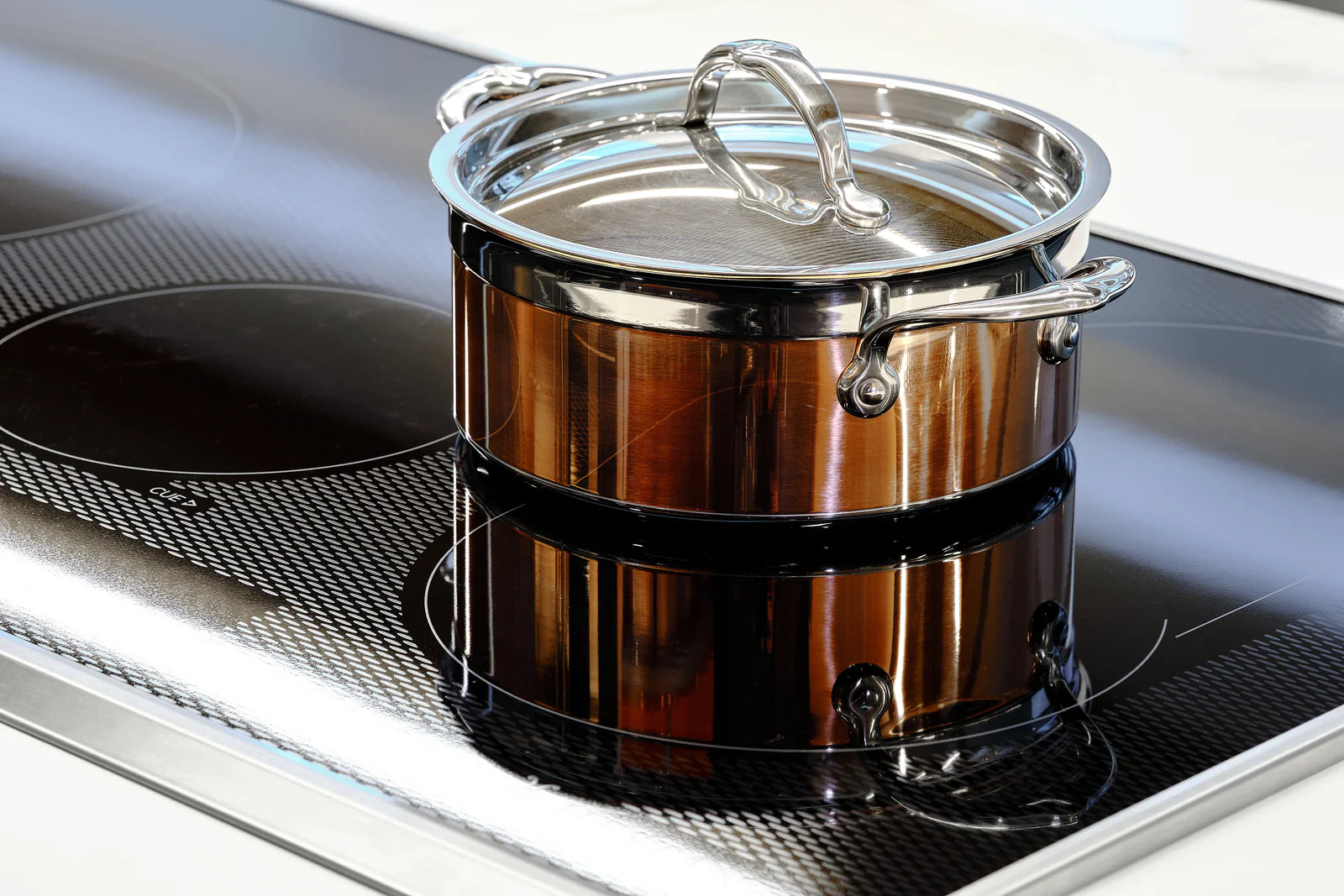 A stainless steel pot with a glass lid sits on an induction cooktop. The cooktop is black with circular heating zones and a textured pattern. The pots shiny surface reflects the light.