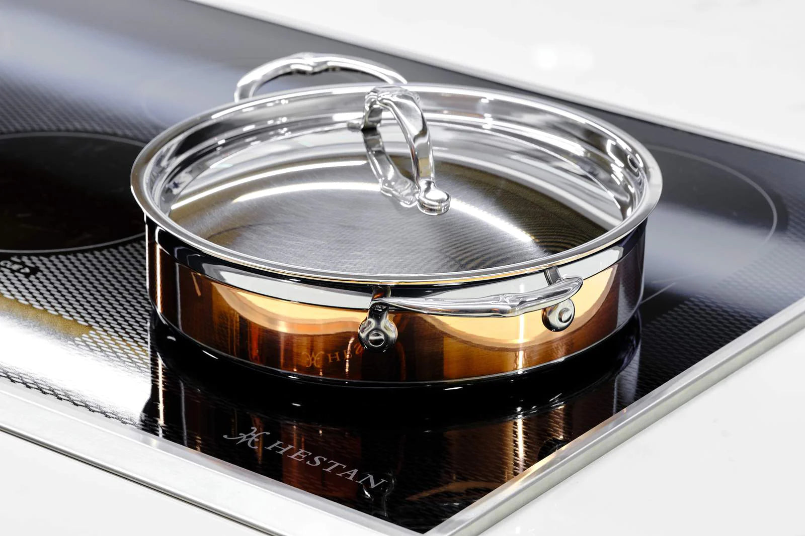 A shiny stainless steel pan with a lid is on an induction cooktop. The pan has a reflective surface, and the cooktop has a glossy finish with visible brand markings.