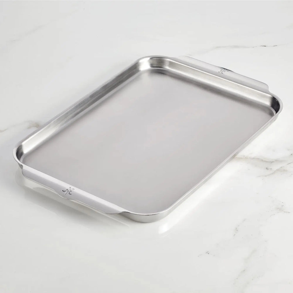 The Hestan OvenBond Tri-ply Half Sheet Pan & Rack (13"x18") with raised edges and two flat handles rests on a white marble surface.