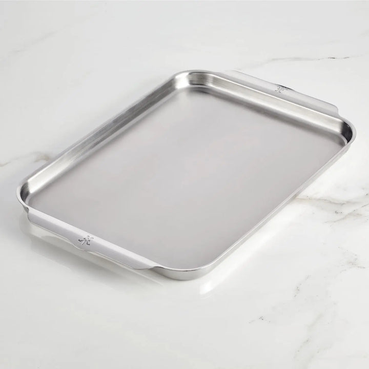 The Hestan OvenBond Tri-ply Half Sheet Pan & Rack (13"x18") with raised edges and two flat handles rests on a white marble surface.