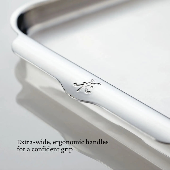 Close-up of the Hestan OvenBond Tri-ply Half Sheet Pan & Rack, 13-in x 18-in, stainless steel with a polished finish and embossed K on the wide, curved handle. Text: Extra-wide, ergonomic handles for a confident grip.