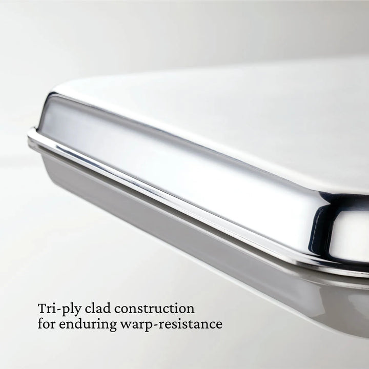 Close-up of a Hestan OvenBond Tri-ply Half Sheet Pan & Rack, 13x18 in, showing its smooth, shiny surface. Text reads: “Tri-ply clad construction for long-lasting warp resistance.”.
