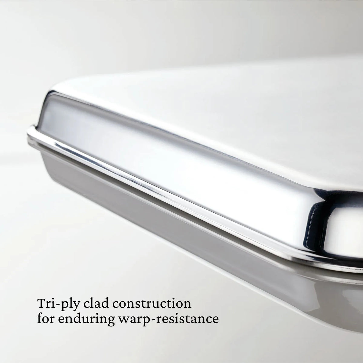 Close-up of a Hestan OvenBond Tri-ply Half Sheet Pan & Rack, 13x18 in, showing its smooth, shiny surface. Text reads: “Tri-ply clad construction for long-lasting warp resistance.”.