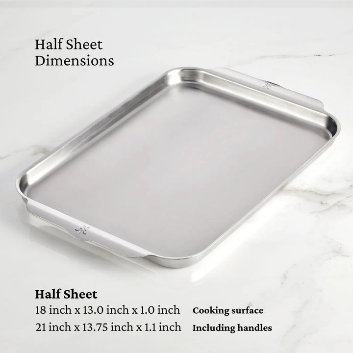 The Hestan OvenBond Tri-ply Half Sheet Pan & Rack, 13-in x 18-in, is shown on a marble surface. Dimensions: 18 x 13 x 1 in (cooking surface), 21 x 13.75 x 1.1 in (with handles)—a versatile roasting sheet pan.