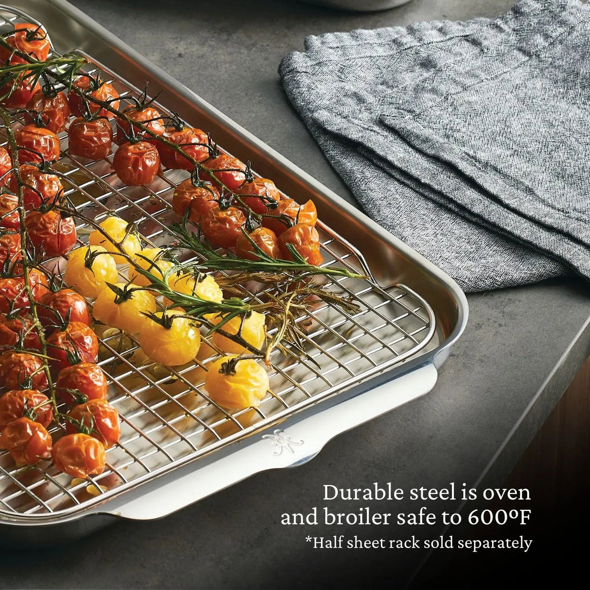 The Hestan OvenBond Tri-ply Half Sheet Pan & Rack (13" x 18") holds roasted red and yellow tomatoes on a counter near folded gray towels. Text reads: “Durable steel is oven and broiler safe to 600°F.”.