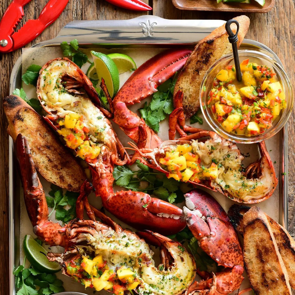 Grilled lobster halves with mango salsa, cilantro, bread slices, and lime wedges are served on a Hestan OvenBond Tri-ply Half Sheet Pan & Rack (13" x 18"); extra salsa in a jar and red-handled lobster crackers sit nearby.