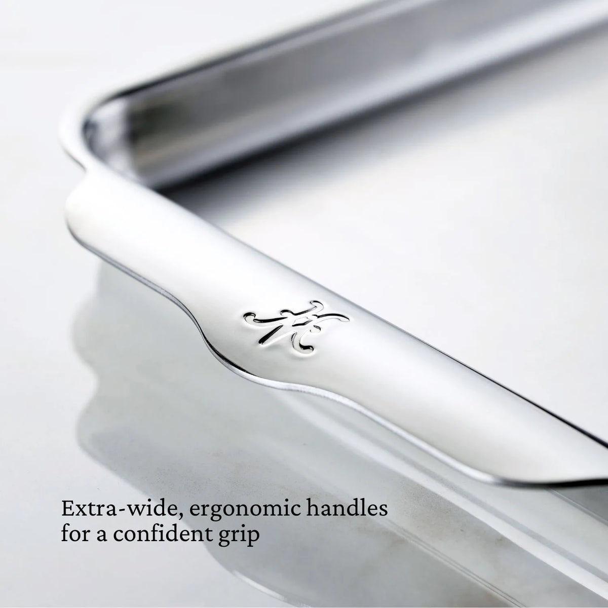 Close-up of a Hestan OvenBond Tri-Ply Quarter Sheet Pan & Rack (9-in x 12-in) from the OvenBond Collection, featuring extra-wide ergonomic handles with engraved logo. Text: Extra-wide, ergonomic handles for a confident grip.