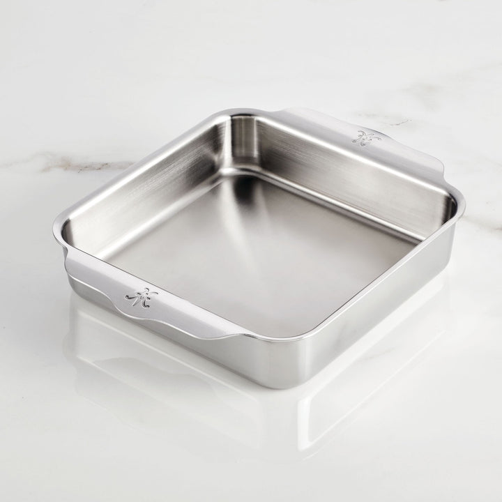 A Hestan OvenBond Tri-ply Square Baker with two handles rests on a white marble surface. The square baker showcases rounded corners and a sleek, polished stainless steel finish.
