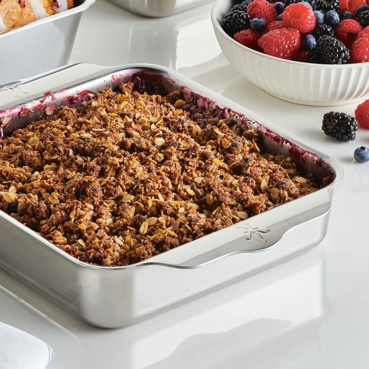 A Hestan OvenBond Tri-ply Square Baker filled with berry crumble sits on a countertop. Beside it, a white bowl contains blueberries, raspberries, blackberries, and strawberries.