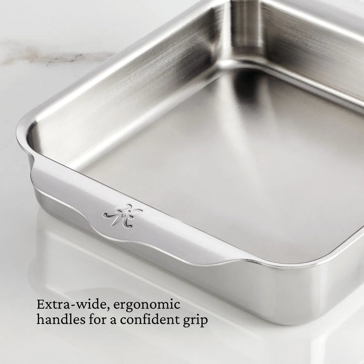 A Hestan OvenBond Tri-ply Square Baker with extra-wide, ergonomic handles sits on a white marble surface. The text reads: Extra-wide, ergonomic handles for a confident grip.