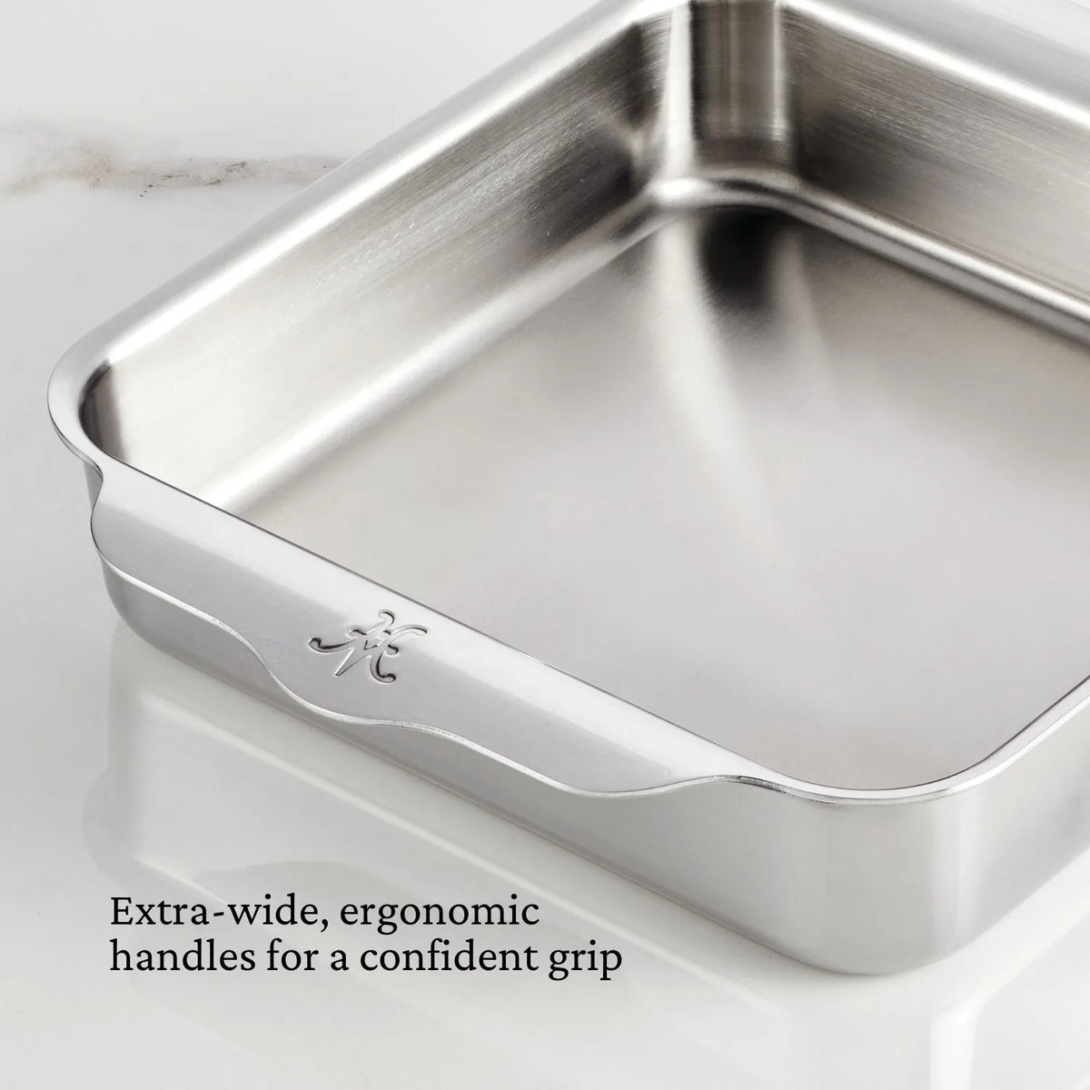 A Hestan OvenBond Tri-ply Square Baker with extra-wide, ergonomic handles sits on a white marble surface. The text reads: Extra-wide, ergonomic handles for a confident grip.