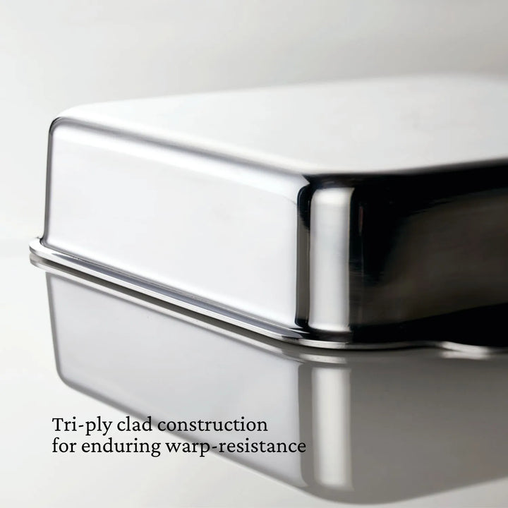 Close-up of a shiny, rectangular Hestan OvenBond Tri-ply Square Baker with smooth edges reflecting on a surface. Text: Hestan OvenBond Tri-ply construction ensures enduring warp-resistance.