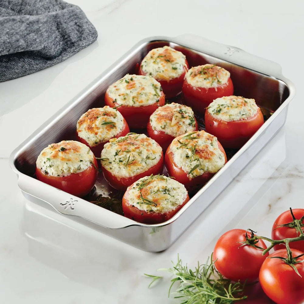 The Hestan OvenBond Tri-ply Rectangular Baker holds nine stuffed tomatoes topped with herbs. The baker sits on a marble countertop, surrounded by vine tomatoes and a gray cloth.