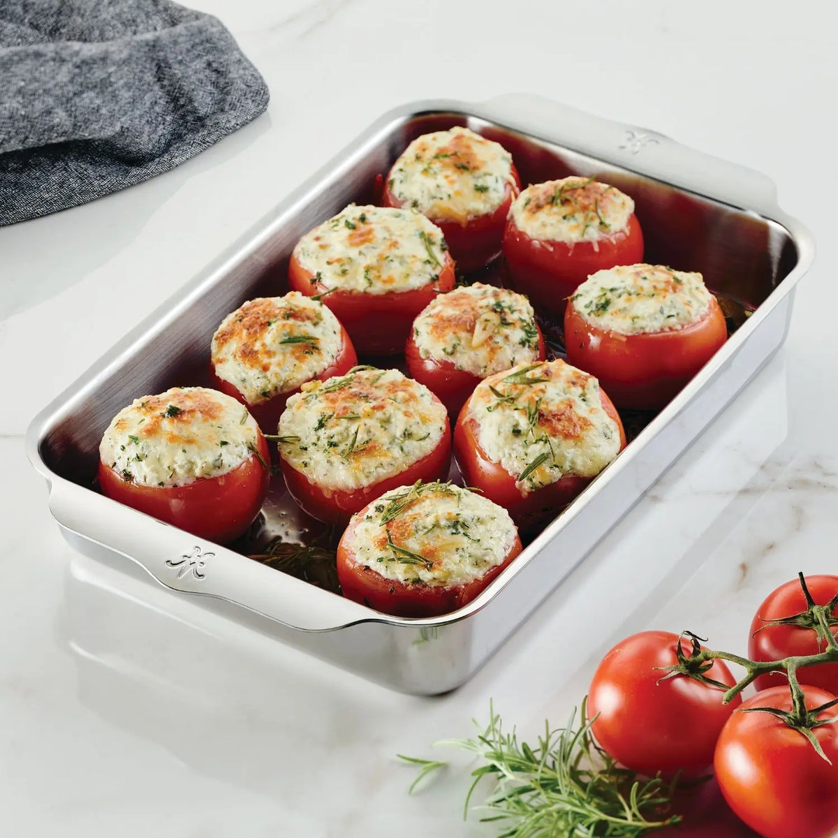 The Hestan OvenBond Tri-ply Rectangular Baker holds nine stuffed tomatoes topped with herbs. The baker sits on a marble countertop, surrounded by vine tomatoes and a gray cloth.