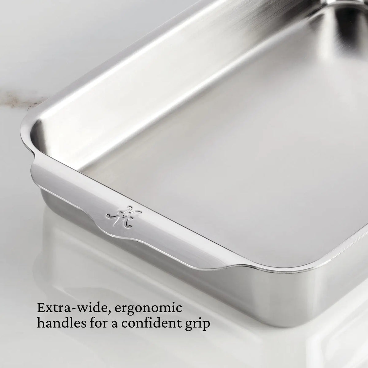 A Hestan OvenBond Tri-ply Rectangular Baker with extra-wide, ergonomic handles and a small engraved logo is shown on a light surface. Caption: Extra-wide, ergonomic handles for a confident grip.
