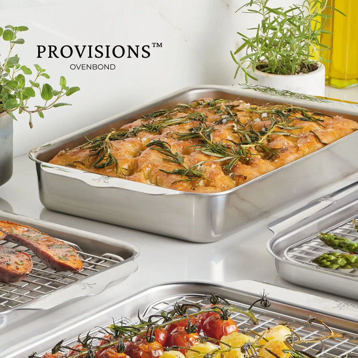 A Hestan OvenBond Tri-ply Rectangular Baker filled with rosemary-topped bread sits on the counter beside pans of roasted vegetables and herbs, with the Hestan OvenBond brand name displayed in the background.
