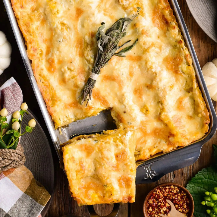 A golden, cheesy lasagna is baked in the Hestan OvenBond Tri-ply Rectangular Baker. One slice is served, a bundle of herbs rests on top, and small bowls of seasonings with a checked napkin sit nearby.