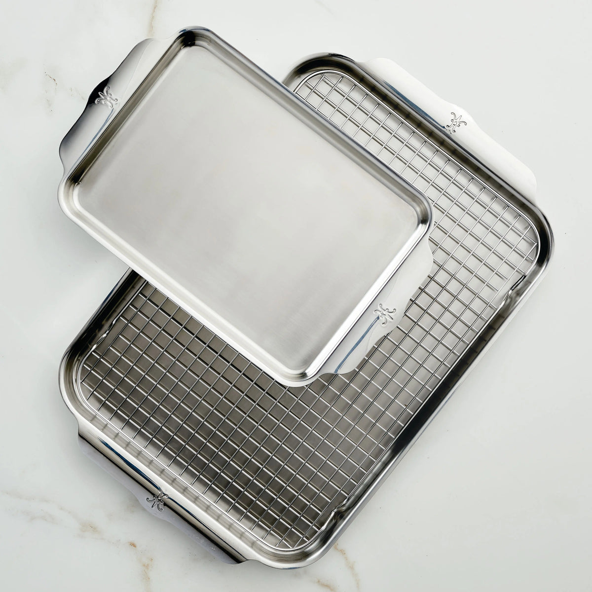 Two Hestan OvenBond Tri-ply 3-Piece Set stainless steel sheet pans rest on a white marble surface, one diagonally atop the other with a wire rack inside. Both have wide, flat handles for easy handling.