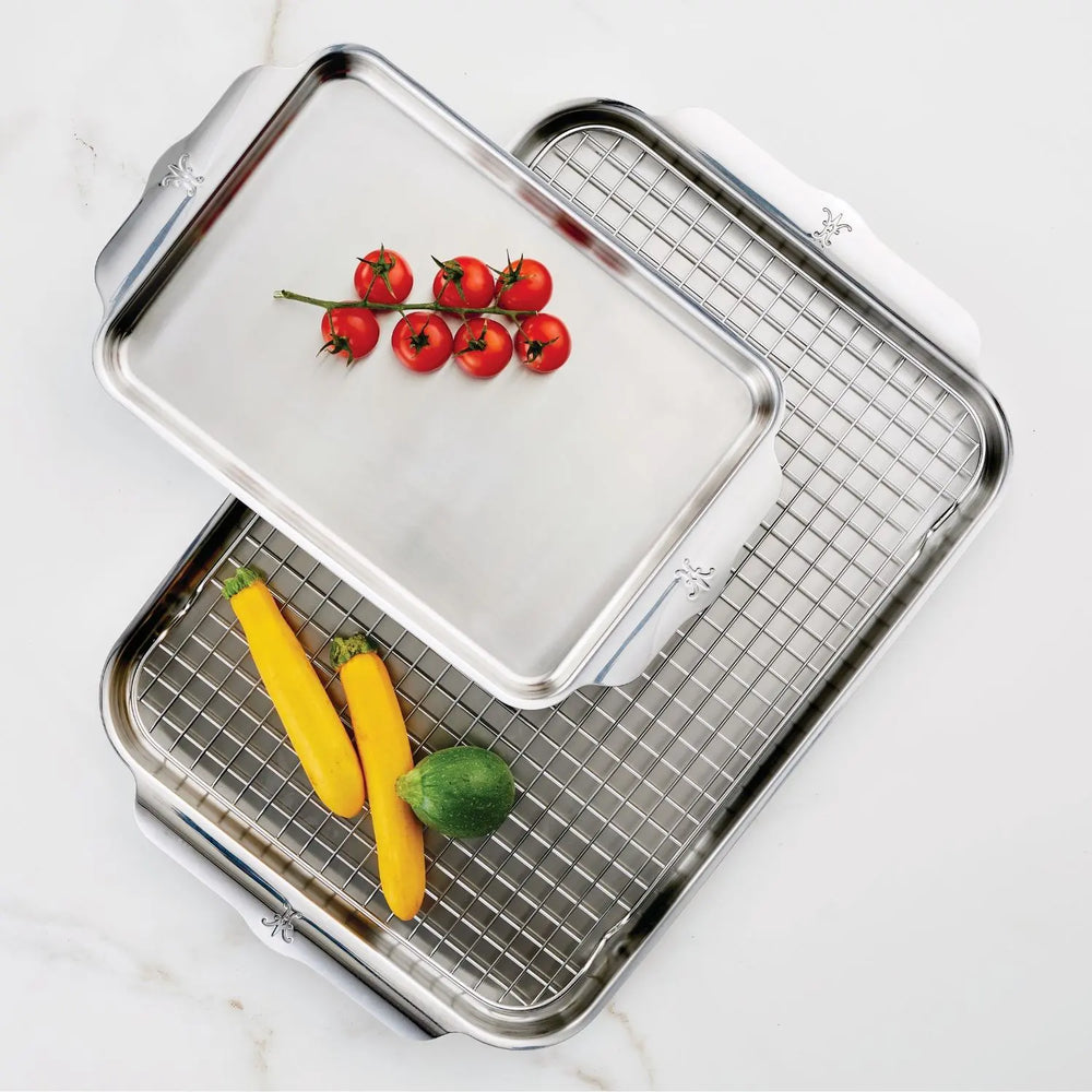 Two stainless steel sheet pans from the Hestan OvenBond Tri-ply 3-Piece Set are shown—one with a rack. The smaller pan contains cherry tomatoes; the larger holds yellow squash and zucchini. Both rest on a white surface.