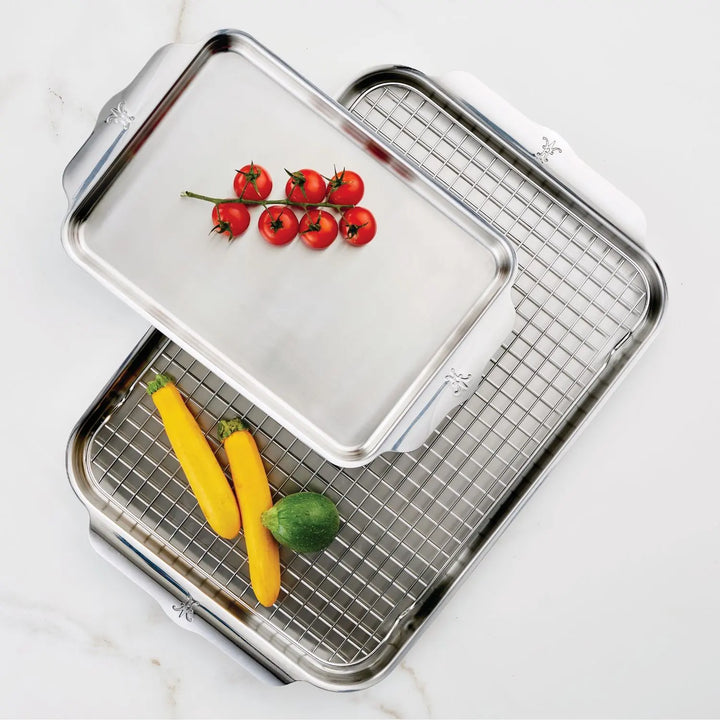 Two stainless steel sheet pans from the Hestan OvenBond Tri-ply 3-Piece Set are shown—one with a rack. The smaller pan contains cherry tomatoes; the larger holds yellow squash and zucchini. Both rest on a white surface.