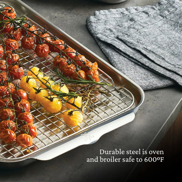 A Hestan OvenBond Tri-ply 5-Piece Set baking sheet with wire rack holds roasted vine tomatoes and rosemary. Folded gray kitchen towels sit nearby. Text: Hestan OvenBond bakeware is oven and broiler safe to 600°F.