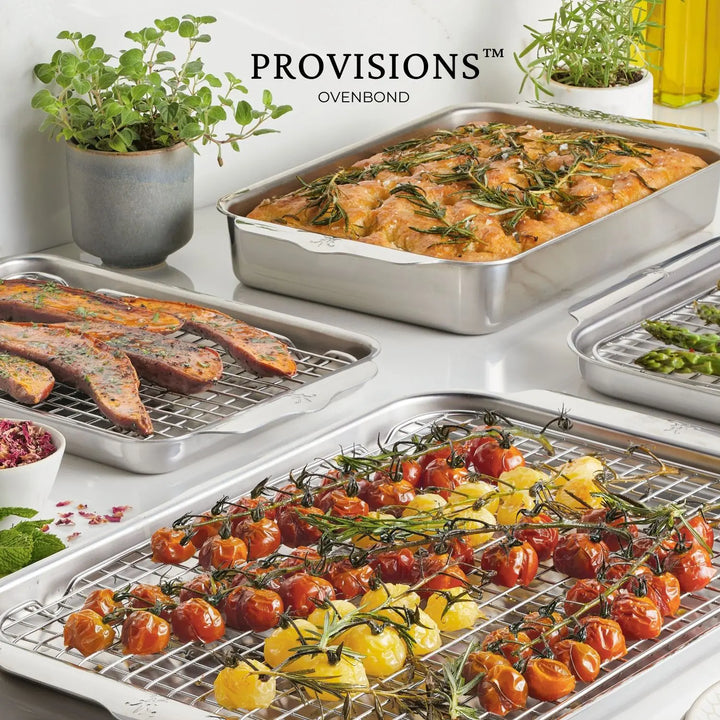 The Hestan OvenBond Tri-ply 3-Piece Set sheet pans are topped with roasted cherry tomatoes, potato wedges, asparagus, and rosemary focaccia bread on a white countertop, with potted herbs in the background.