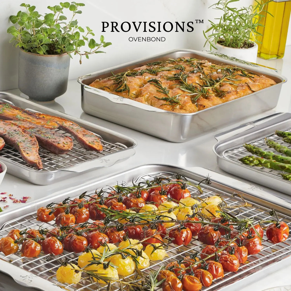 A kitchen countertop showcases the Hestan OvenBond Tri-ply 5-Piece Set, filled with roasted cherry tomatoes, rosemary focaccia, asparagus, and carrots. Potted herbs and olive oil sit in the background.
