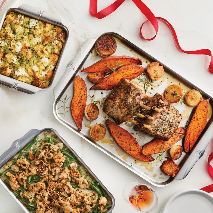 A roasted rack of pork with sweet potato wedges, herbed cheese stuffing, and crispy onion green beans are baked in the Hestan OvenBond Tri-ply 5-Piece Set. Nearby are a glass of rosé and red ribbon for a festive touch.