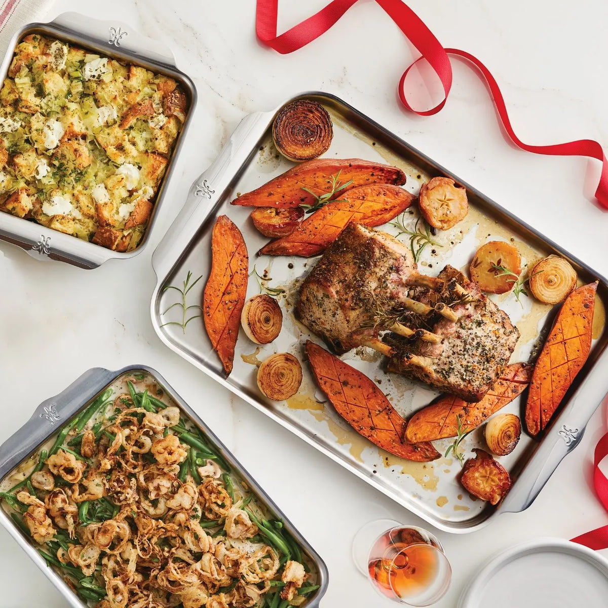 A roasted rack of pork with sweet potato wedges, herbed cheese stuffing, and crispy onion green beans are baked in the Hestan OvenBond Tri-ply 5-Piece Set. Nearby are a glass of rosé and red ribbon for a festive touch.