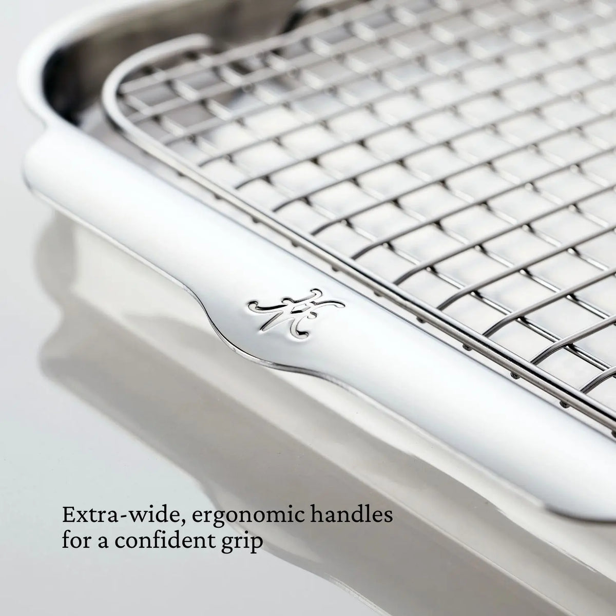 Close-up of a Hestan OvenBond Tri-ply 5-Piece Set cooling rack featuring extra-wide, ergonomic handles engraved with a decorative letter. Text reads: Extra-wide, ergonomic handles for a confident grip.