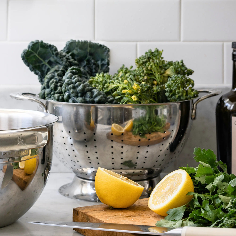 The Hestan Provisions Stainless Steel Colander, dishwasher safe and designed for superior drainage, overflows with leafy greens on the kitchen counter amid a cut lemon, chopped greens, a knife, a mixing bowl, and a bottle—perfect for fresh meal prep.