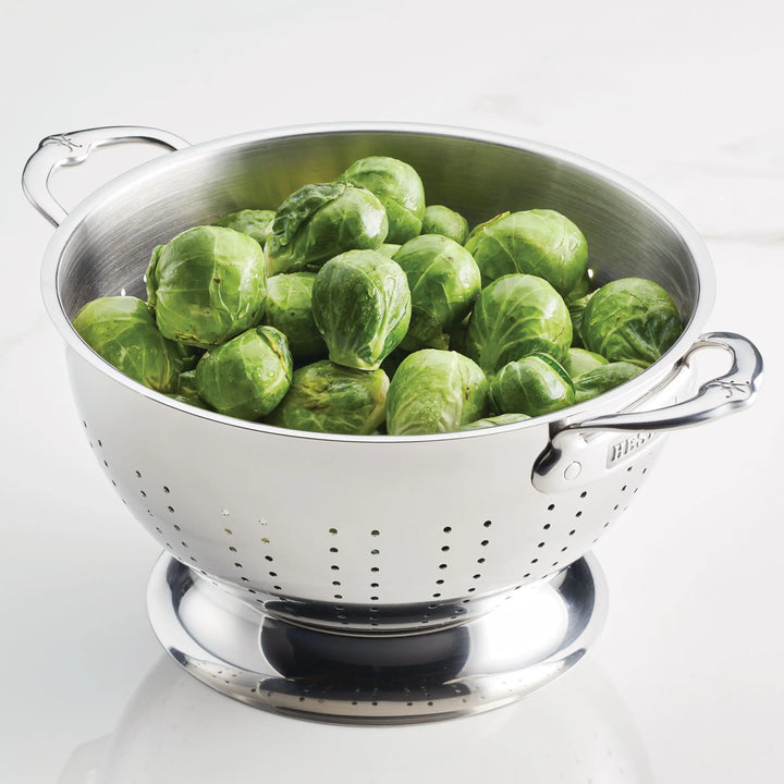 A Hestan Provisions Stainless Steel Colander with excellent drainage holds fresh Brussels sprouts and rests on a white surface.