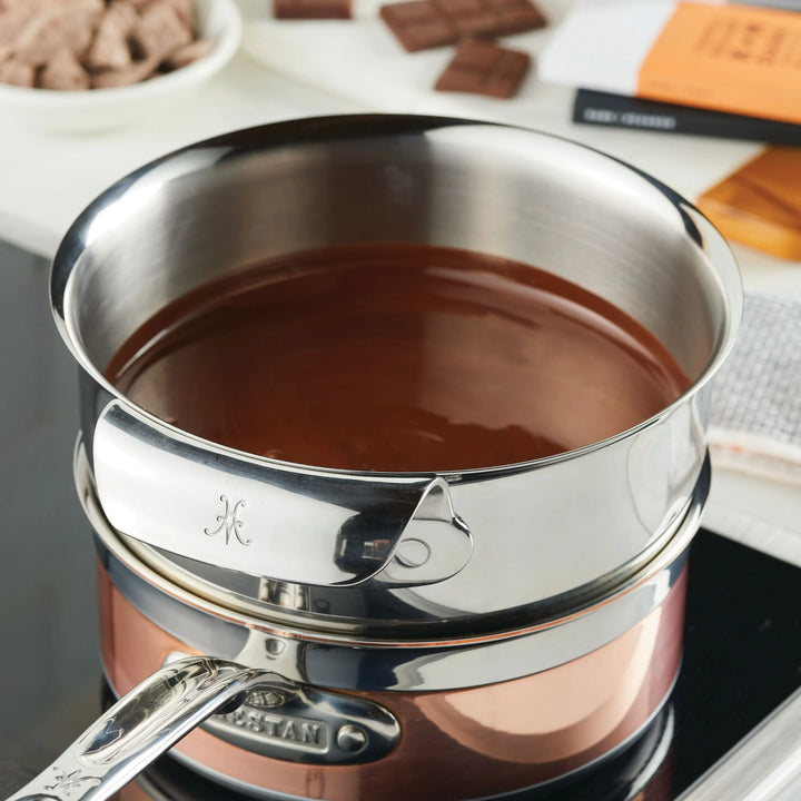 A metal saucepan melts chocolate on a stove, with Hestan Provisions Stainless Steel Mixing Bowl Set (3-Piece) nearby. Chocolate bars and a white bowl with chocolate chunks appear in the background as part of the double boiler setup.