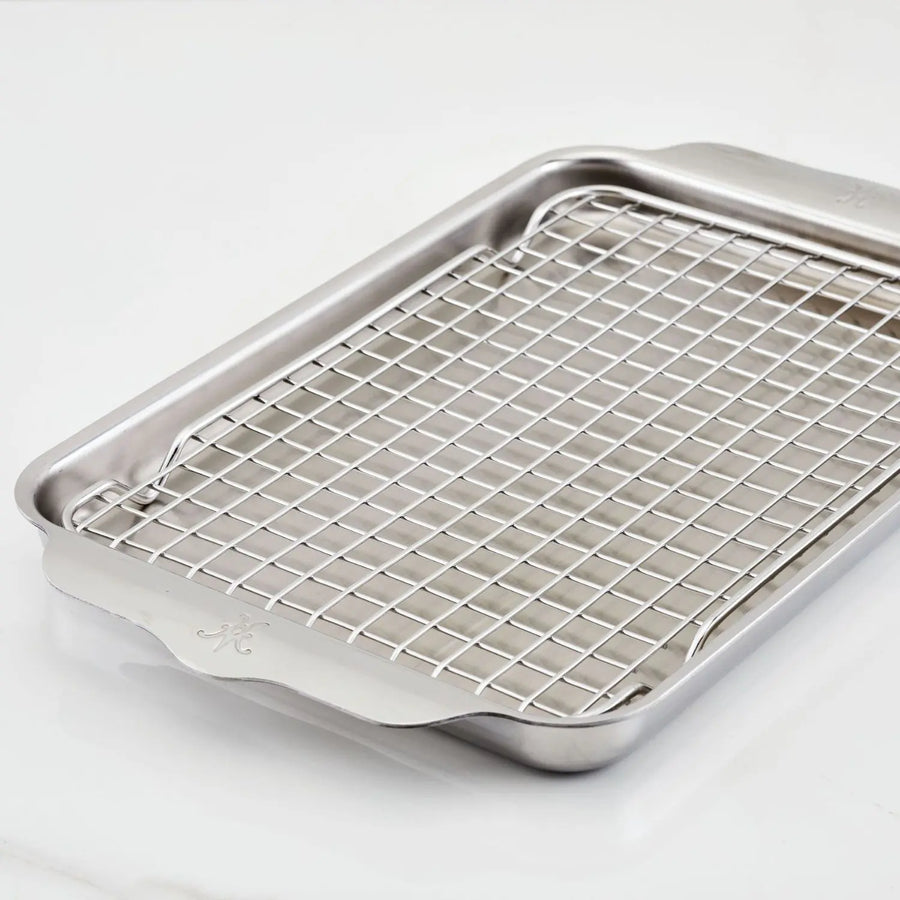 The Hestan OvenBond Tri-Ply Quarter Sheet Pan & Rack (9-in x 12-in) from the OvenBond Collection is set on a white marble surface, featuring side handles, a slightly raised edge, and a matching wire cooling rack inside.