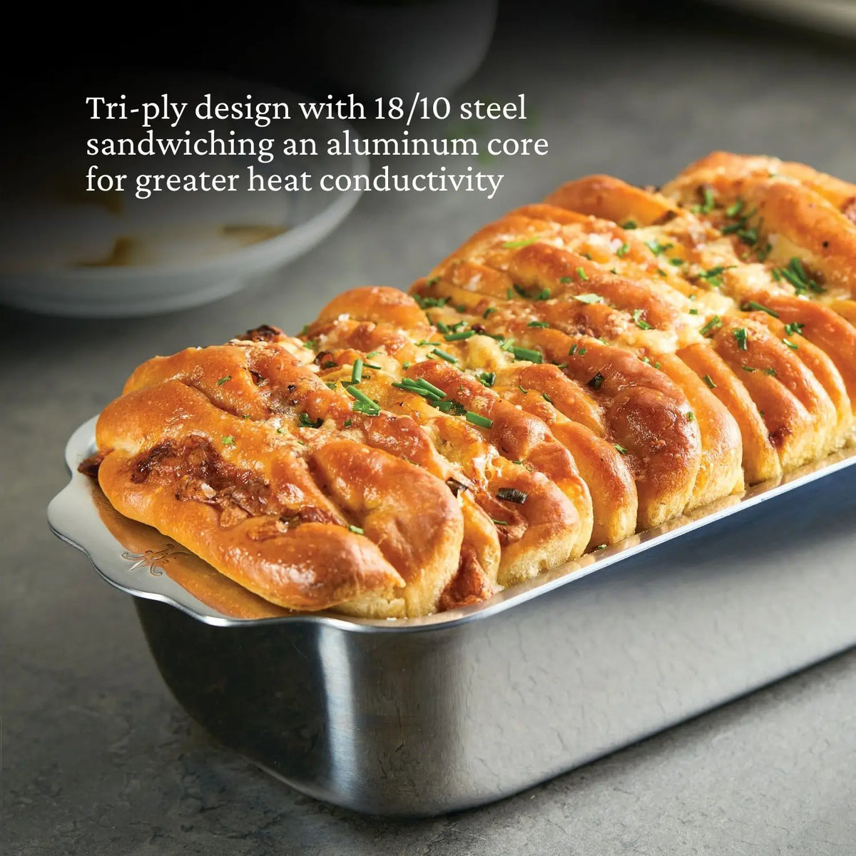 A golden-brown, pull-apart bread with herbs sits in the Hestan OvenBond Tri-ply Stainless Steel 1-Pound Loaf Pan, featuring durable 18/10 steel and an aluminum core for even heating. White text spotlights its oven-safe tri-ply design.