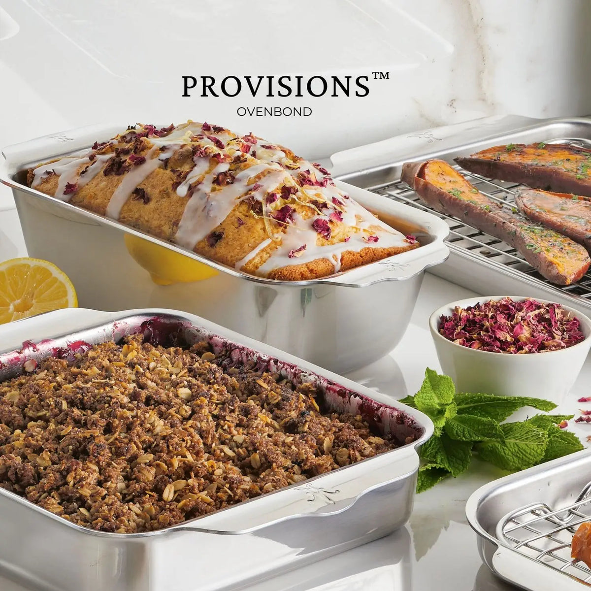 Three Hestan OvenBond Tri-Ply 3-Piece Baking Set pans hold a glazed loaf cake with rose petals, salmon fillets, and fruit crumble, accented by lemon halves, mint leaves, and dried rose petals. The text reads: Hestan OvenBond.