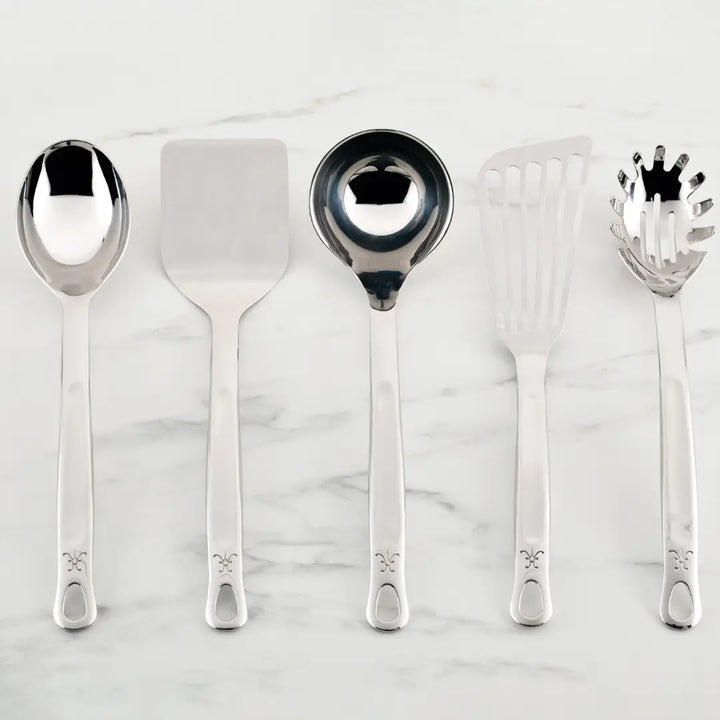 A set of five Hestan Provisions Essential Chefs Tools, including a serving spoon, flat spatula, ladle, slotted spatula, and spaghetti server, elegantly rest on a white marble surface. Each piece boasts a handle with a decorative emblem.