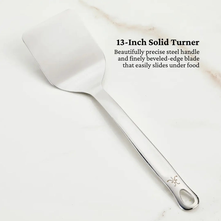 The Deluxe Chefs Tool Set by Hestan Provisions includes a 13-inch stainless steel solid turner spatula with a flat, beveled-edge blade for easy food handling and a handle elegantly adorned with a fleur-de-lis design.