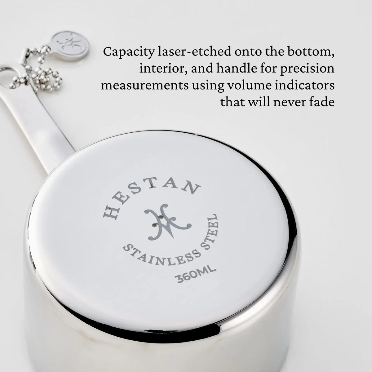 A stainless steel cup from the Hestan Provisions® Odd-Size Measuring Cups Set, 3-Piece is shown upside down, revealing the bottom with the Hestan logo, STAINLESS STEEL, and 360ML laser-etched markings. A keychain and tag are attached to the handle, highlighting its inclusion in the premium stainless steel measuring cup set series.
