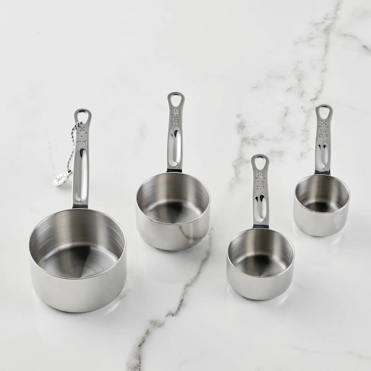 Four stainless steel measuring cups of different sizes are arranged on a white marble surface. The Standard Size Measuring Cups Set, 4-Piece from Hestan Provisions® features handles marked with each cup’s measurement. The largest cup is positioned on the left, while the smallest sits on the right.