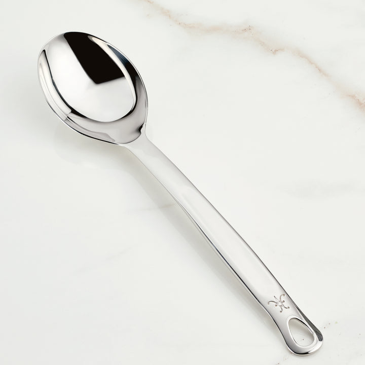 The Hestan Provisions Deep Solid Spoon, 13-inch, made from 18/10 stainless steel with a smooth rounded bowl and an engraved design on the handle, elegantly rests on a subtly marbled white surface.