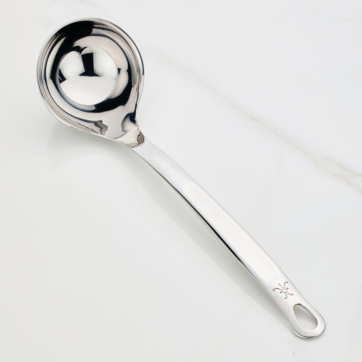 A Hestan Provisions Ladle, 5-ounce, with a shiny finish is elegantly placed on a white surface. It features a deep, rounded bowl and a long handle with a hole at the end for convenient hanging.