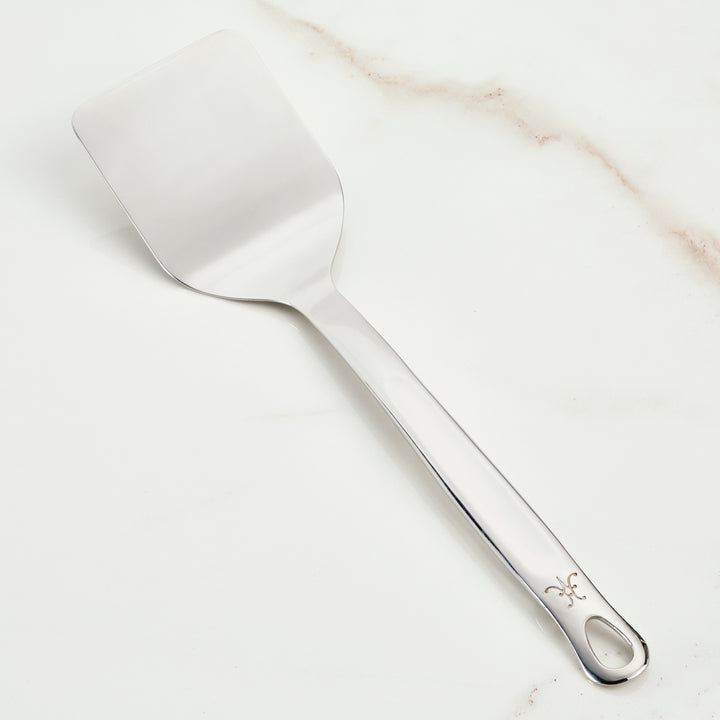 The Hestan Provisions Solid Turner (13-inch), crafted from premium 18/10 stainless steel, boasts a flat, rectangular blade and a long handle adorned with a decorative symbol. Elegantly displayed on white marble, its perfect for Michelin-starred chefs.
