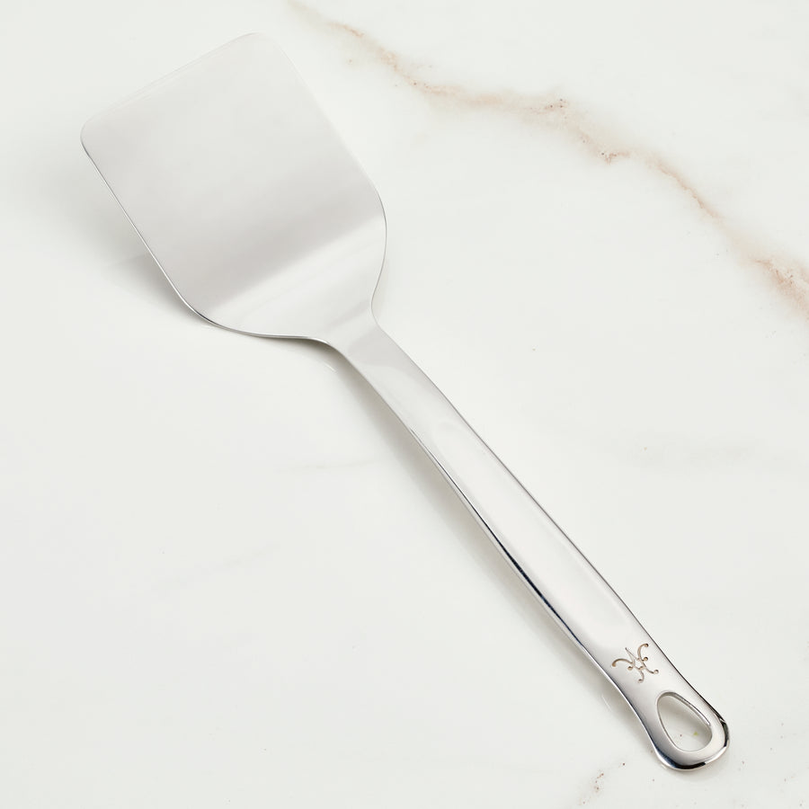 The Hestan Provisions Solid Turner (13-inch), crafted from premium 18/10 stainless steel, boasts a flat, rectangular blade and a long handle adorned with a decorative symbol. Elegantly displayed on white marble, its perfect for Michelin-starred chefs.