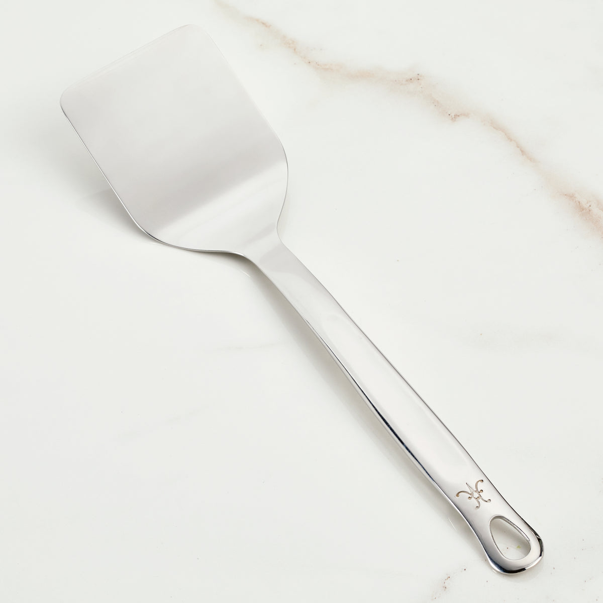 The Hestan Provisions Solid Turner (13-inch), crafted from premium 18/10 stainless steel, boasts a flat, rectangular blade and a long handle adorned with a decorative symbol. Elegantly displayed on white marble, its perfect for Michelin-starred chefs.
